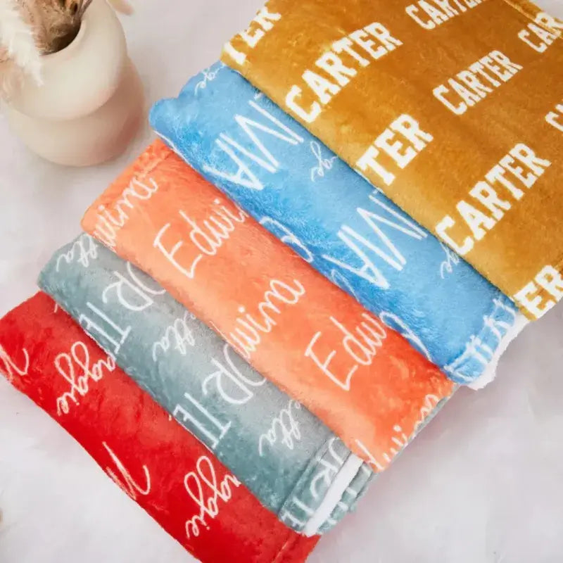 Cinq plaids personnalisés pliés dans différentes couleurs - rouge, gris, corail, bleu et moutarde - avec textes personnalisés en blanc, avec une tasse en arrière-plan