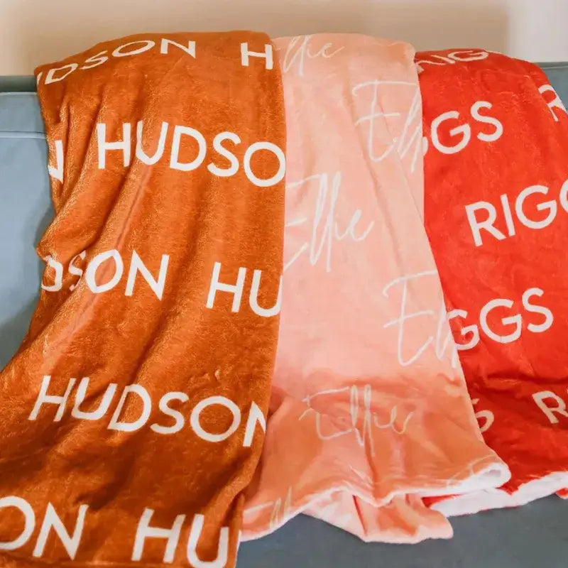 Trois plaids personnalisés empilés - orange Hudson, rose poudré avec signature, et rouge avec texte blanc - affichant les options de couleurs disponibles
